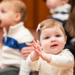 Smiling baby claps hands while sitting with children and adults during a community gathering at Temple Emanu-El.