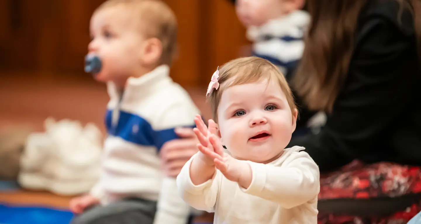 Smiling baby claps hands while sitting with children and adults during a community gathering at Temple Emanu-El.