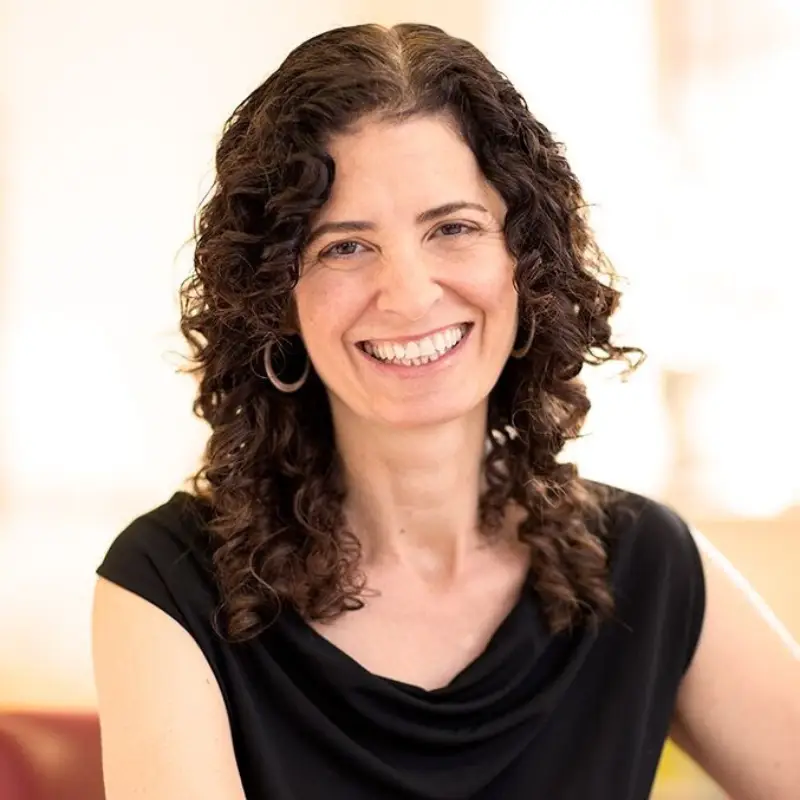 Woman with curly brown hair, in a black top, smiling warmly at the camera in a softly lit indoor setting at Temple Emanu-El.