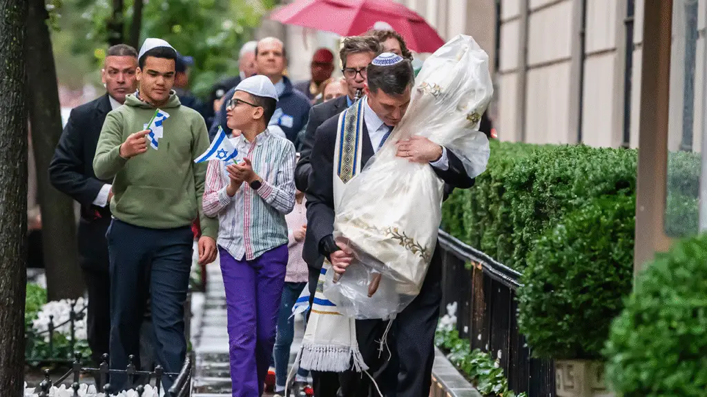 Congregants walk together in the rain outside Temple Emanu-El; one man reverently carries a Torah scroll wrapped in a white cloth.