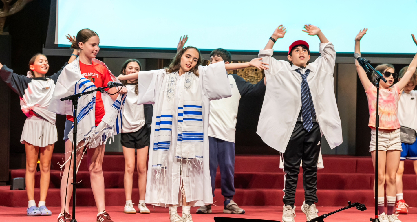 Children on stage with arms raised; two wear tallitot while others are in casual attire, joyfully performing at Temple Emanu-El.