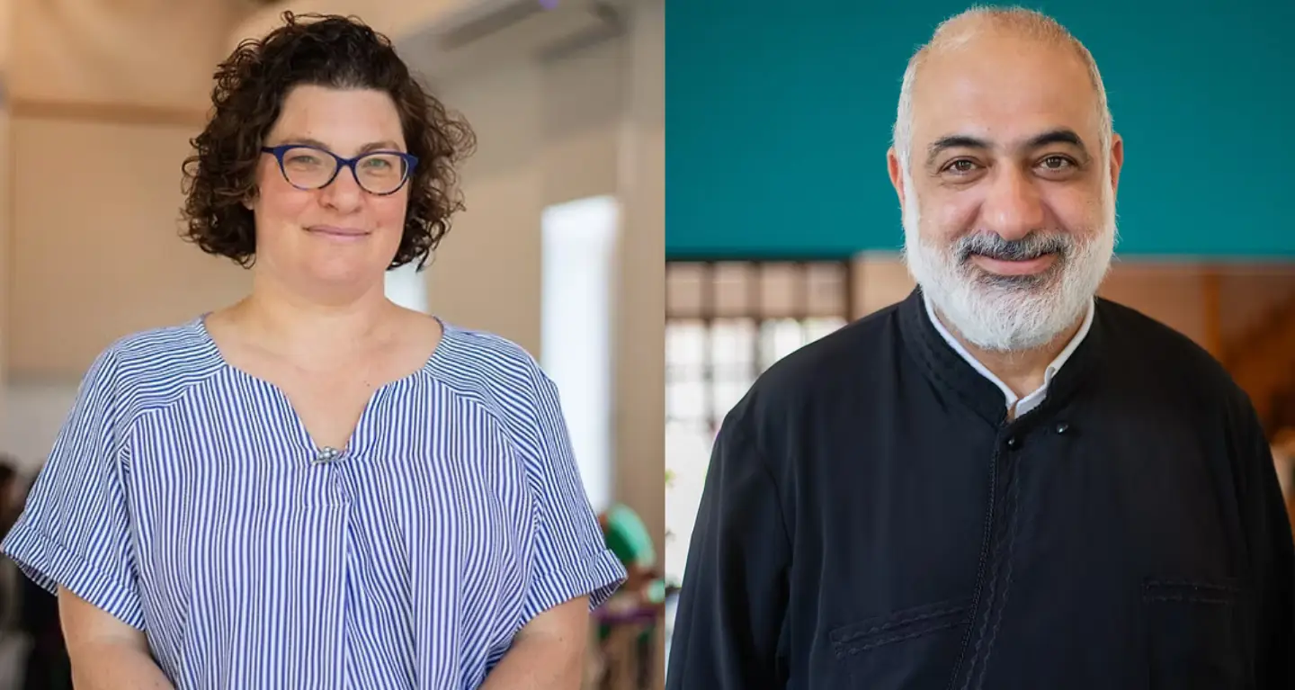 Two smiling adults indoors at Temple Emanu-El, one woman in glasses and stripes, one bearded man in a dark top.