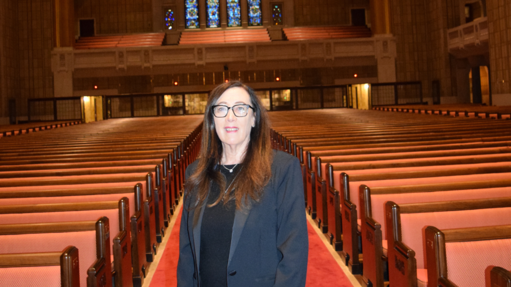Woman with long brown hair and glasses stands in an empty synagogue with wooden pews and stained glass windows.