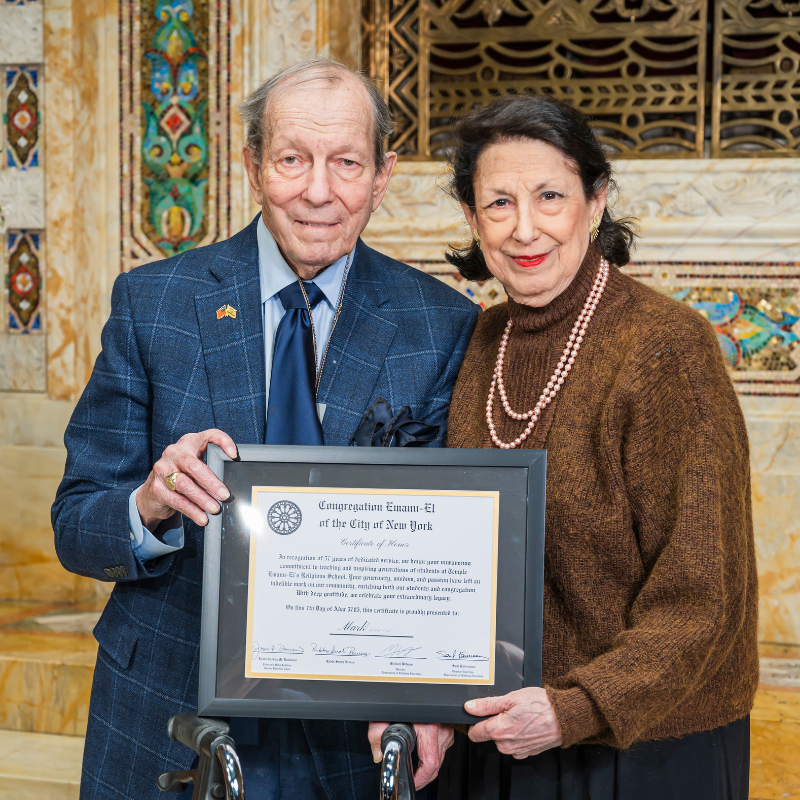 An older man and woman stand together inside Temple Emanu-El, smiling and holding a framed certificate for the camera.