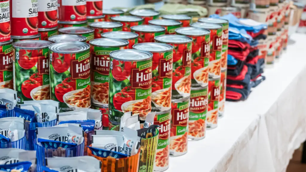 Rows of Hunts pasta sauce cans and assorted packaged foods arranged on a table at Temple Emanu-El for a food drive.