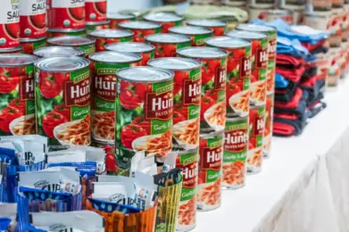 Rows of Hunts pasta sauce cans and assorted packaged foods arranged on a table at Temple Emanu-El for a food drive.