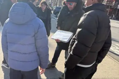 A group of visitors stand on the city sidewalk outside looking at a map held by one person in a black jacket and beanie.