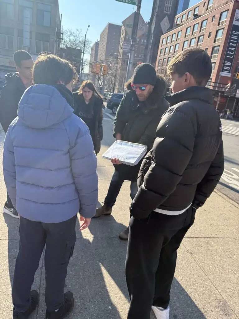 A group of visitors stand on the city sidewalk outside looking at a map held by one person in a black jacket and beanie.