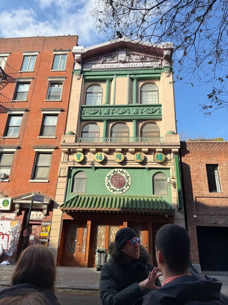 People stand in front of a grand beige and green building with Hebrew characters on the facade.