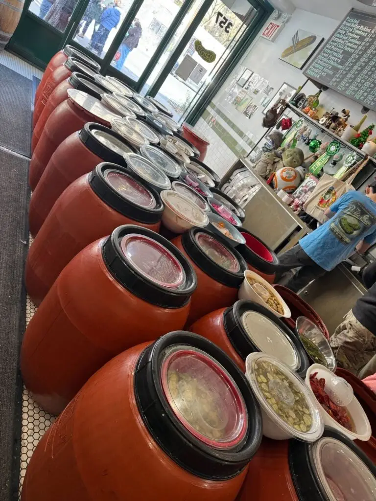 Red barrels brimming with assorted pickles are neatly arranged along a well-lit aisle in a welcoming market setting.