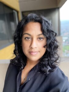 Woman with shoulder-length curly hair, in a black blouse, stands on a balcony with a softly blurred background.
