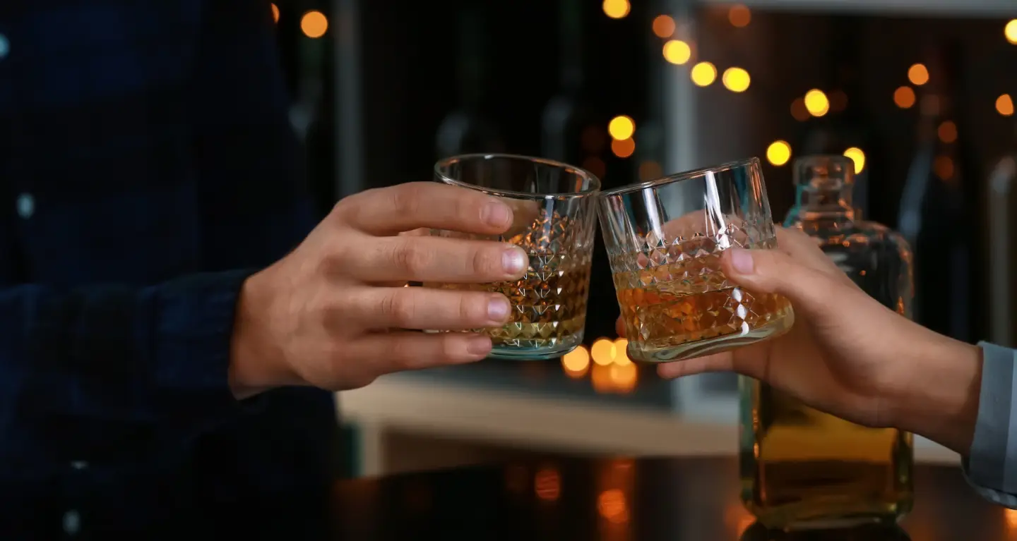 Two people sharing a toast with drinks in hand, with a bottle and soft, warm lighting in the background.