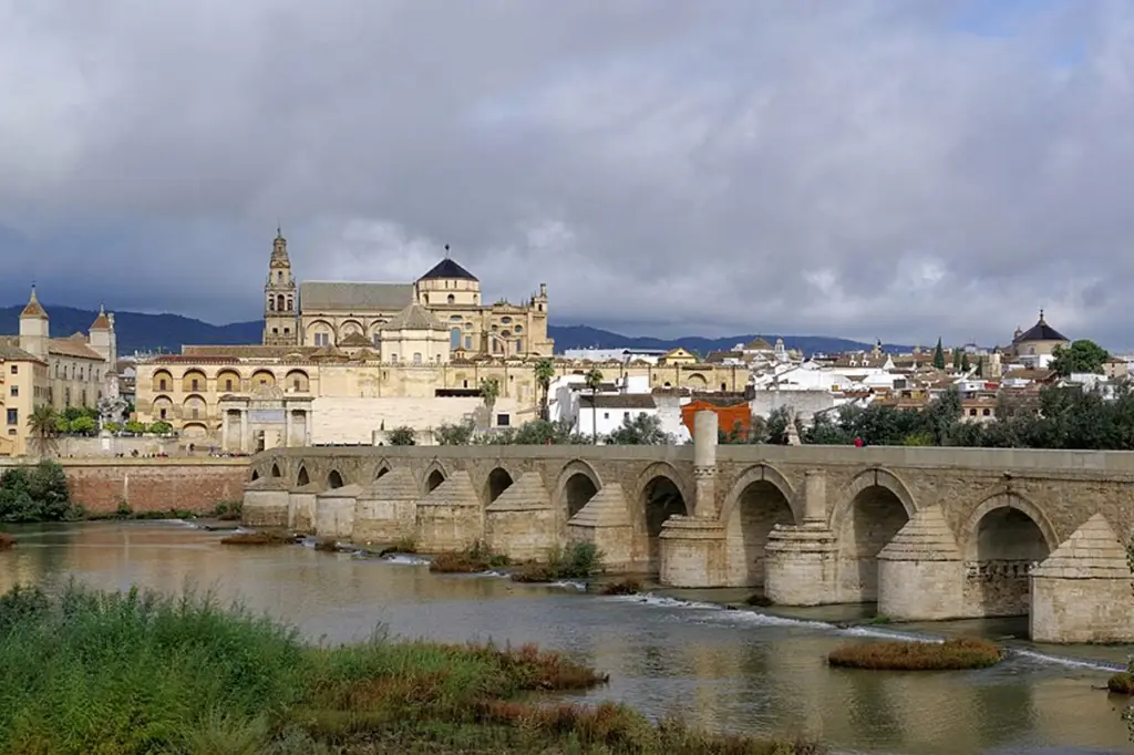 Historic stone bridge in Cordoba, Spain, crosses a river with buildings in the background beneath cloudy skies.