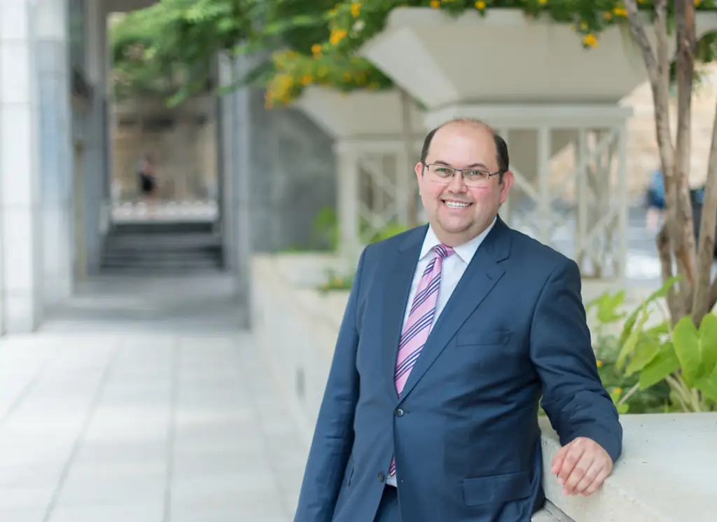 A man in a suit and glasses leans on a ledge outdoors smiling warmly at the camera.