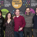Seven people smiling, standing together in front of a colorful Friday Night Hub backdrop at Temple Emanu-El in New York City.