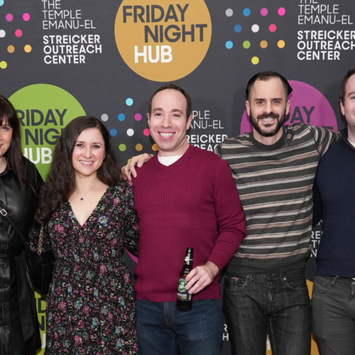 Seven people smiling, standing together in front of a colorful Friday Night Hub backdrop at Temple Emanu-El in New York City.