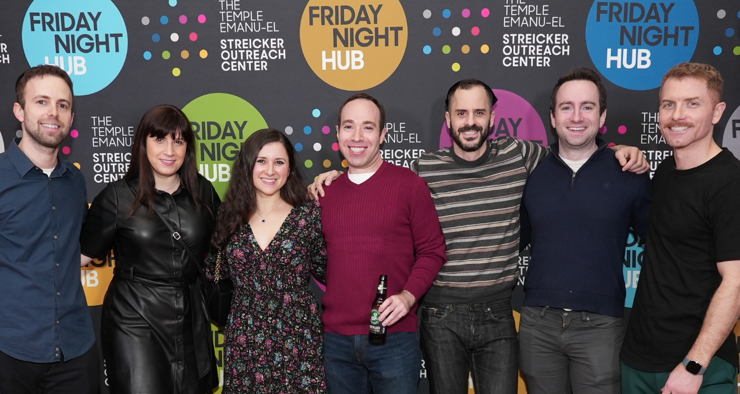 Seven people smiling, standing together in front of a colorful Friday Night Hub backdrop at Temple Emanu-El in New York City.