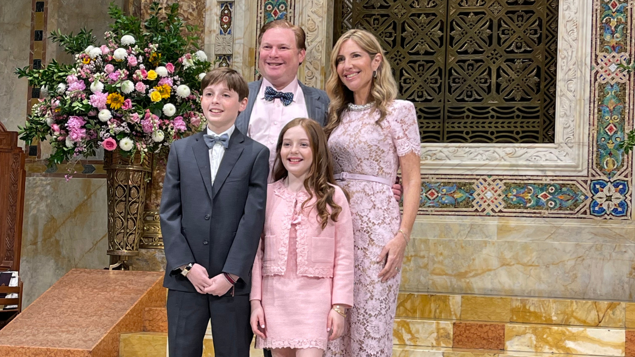 A family of four, dressed formally, smiles and poses together in Temple Emanu-El with elegant décor and floral arrangements.