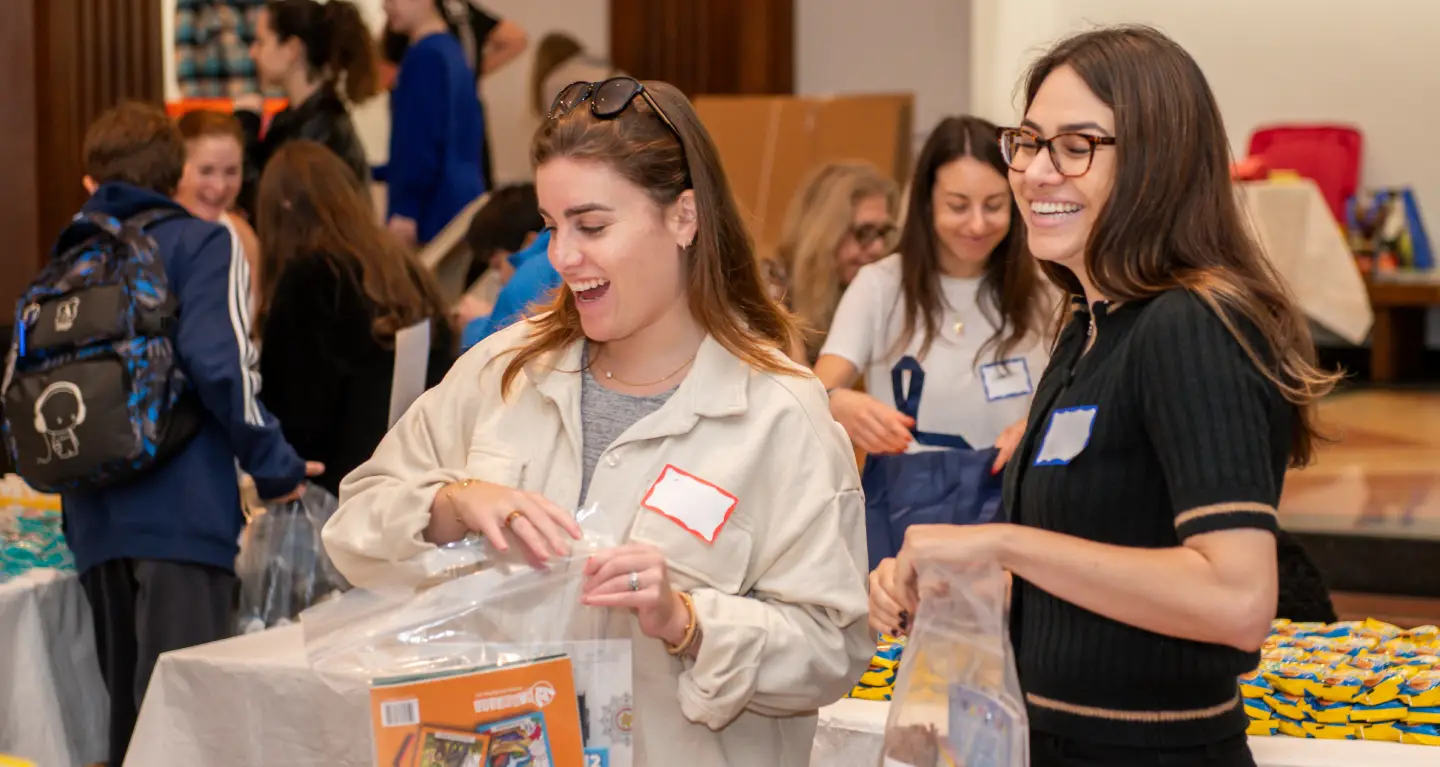 Two women smiling as they pack items into bags at a Temple Emanu-El community volunteer event, with others in the background.