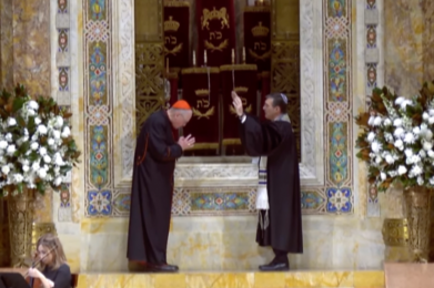 A visiting cardinal bows as a rabbi offers a blessing at Temple Emanu-El’s bimah, flanked by white flower arrangements.