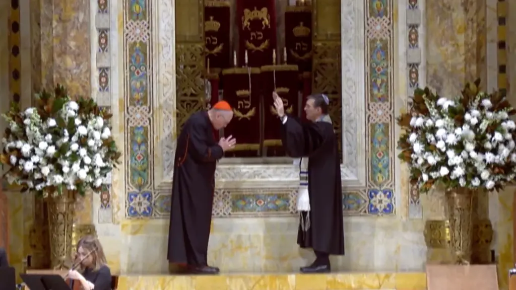 A visiting cardinal bows as a rabbi offers a blessing at Temple Emanu-El’s bimah, flanked by white flower arrangements.