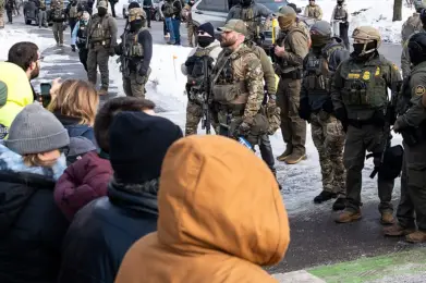 ICE officers stand on a snowy street facing a group of people during a public demonstration.