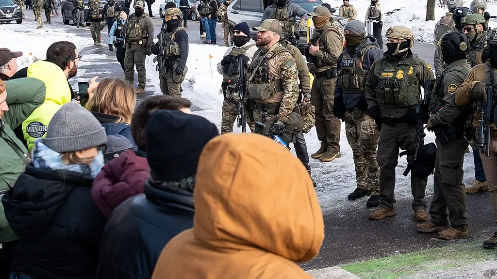 ICE officers stand on a snowy street facing a group of people during a public demonstration.