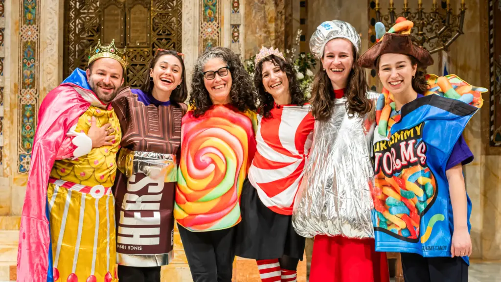 Six people wearing colorful candy and snack-themed costumes share smiles indoors at Temple Emanu-El.
