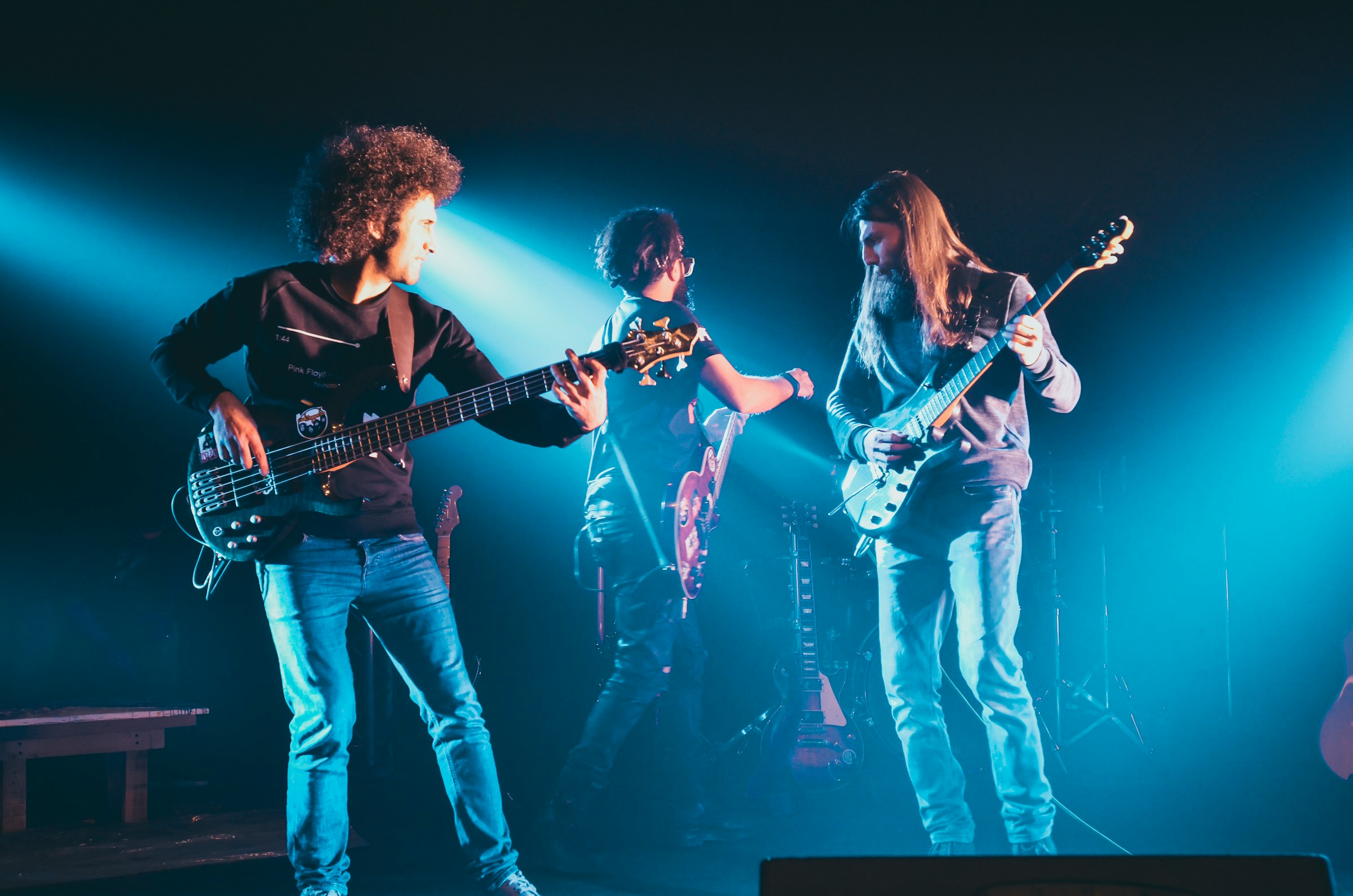 Three musicians perform with electric guitars onstage, illuminated by blue lights, at a Temple Emanu-El community event.