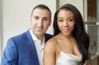 A man in a blue suit and a woman in a white dress smile together by a window.