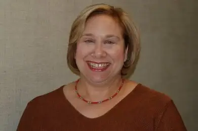 A smiling woman in a brown sweater and red beaded necklace stands in front of a neutral wall.