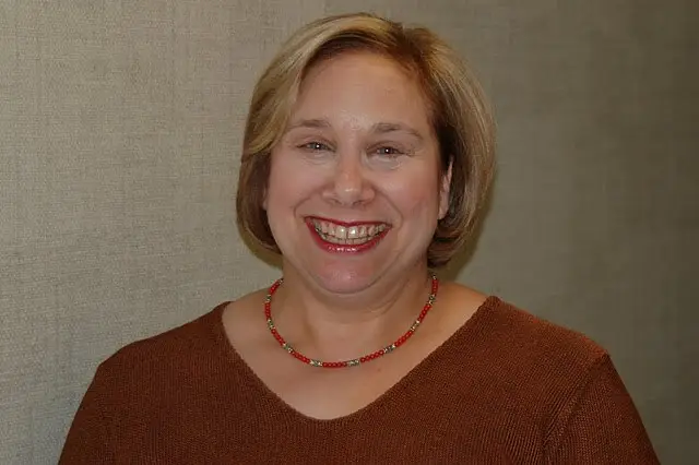 A smiling woman in a brown sweater and red beaded necklace stands in front of a neutral wall.