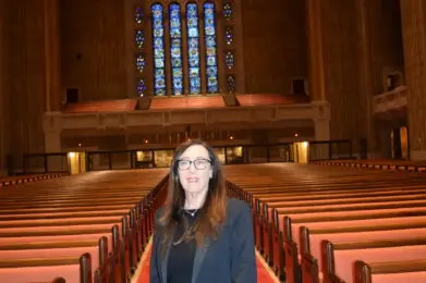 A woman in black stands on a red carpet inside a large synagogue with ornate stained glass windows.