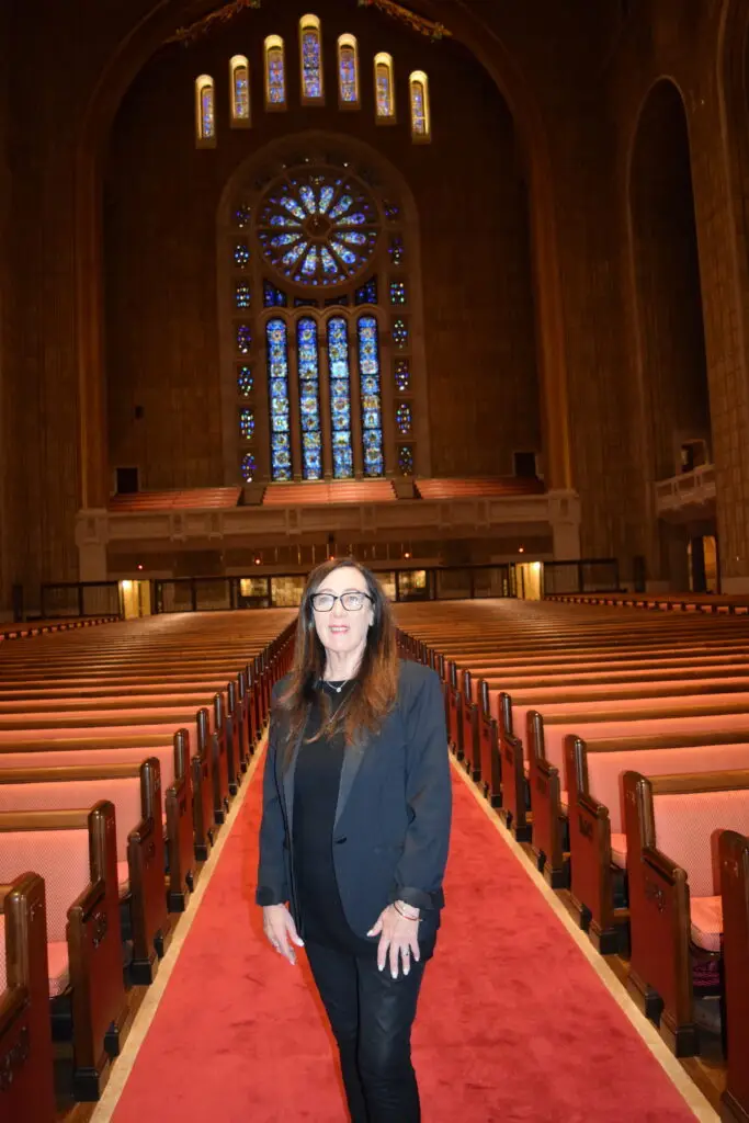 A woman in black stands on a red carpet inside a large synagogue with ornate stained glass windows.