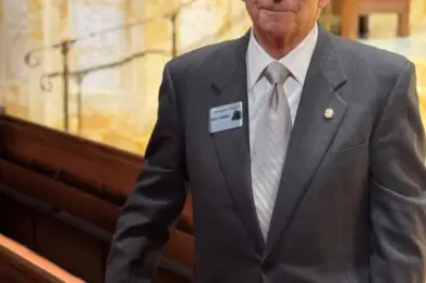 Older man in a gray suit stands in a synagogue pew at Temple Emanu-El, wearing glasses and a name badge, smiling slightly.
