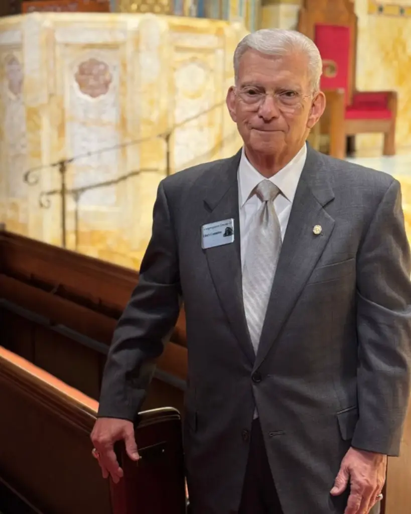 Older man in a gray suit stands in a synagogue pew at Temple Emanu-El, wearing glasses and a name badge, smiling slightly.