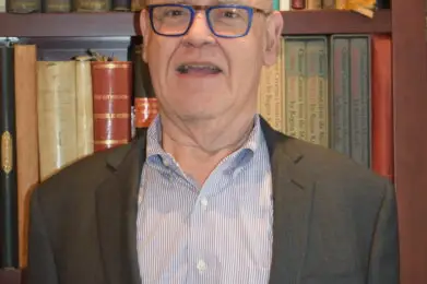 An older man in glasses and a suit stands smiling in front of a bookshelf, creating a welcoming atmosphere at Temple Emanu-El.