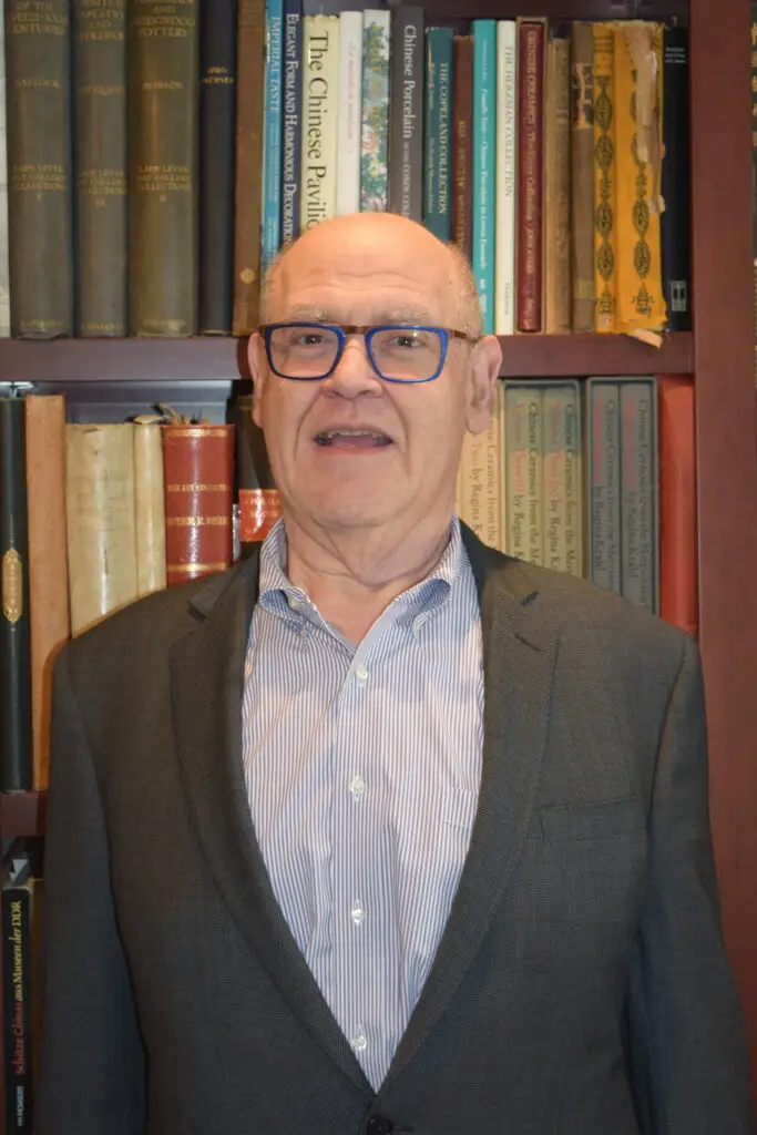 An older man in glasses and a suit stands smiling in front of a bookshelf, creating a welcoming atmosphere at Temple Emanu-El.