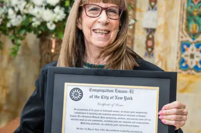 Smiling woman with glasses holds a framed certificate inside the ornate sanctuary of Temple Emanu-El, flowers in the background.