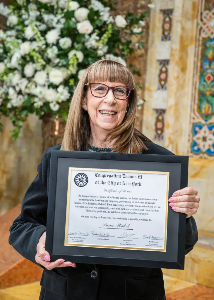 Smiling woman with glasses holds a framed certificate inside the ornate sanctuary of Temple Emanu-El, flowers in the background.