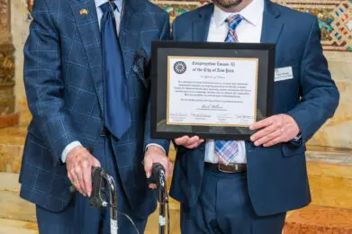 Two men in suits smile on the marble steps of Temple Emanu-El, one proudly holding a framed certificate.