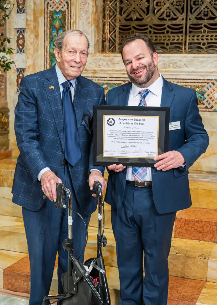 Two men in suits smile on the marble steps of Temple Emanu-El, one proudly holding a framed certificate.