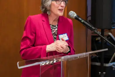 A woman in a pink blazer speaks at the podium with a microphone during an event at Temple Emanu-El in New York City.
