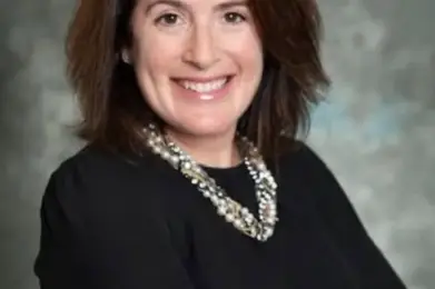 Smiling woman with shoulder-length brown hair, in a black top and beaded necklace, standing with arms crossed at Temple Emanu-El.