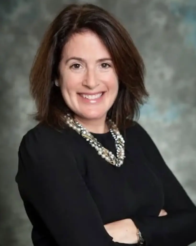 Smiling woman with shoulder-length brown hair, in a black top and beaded necklace, standing with arms crossed at Temple Emanu-El.