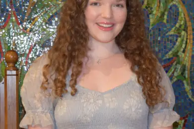 A young woman with curly hair in a light dress smiles while standing before a vibrant mosaic wall at Temple Emanu-El.