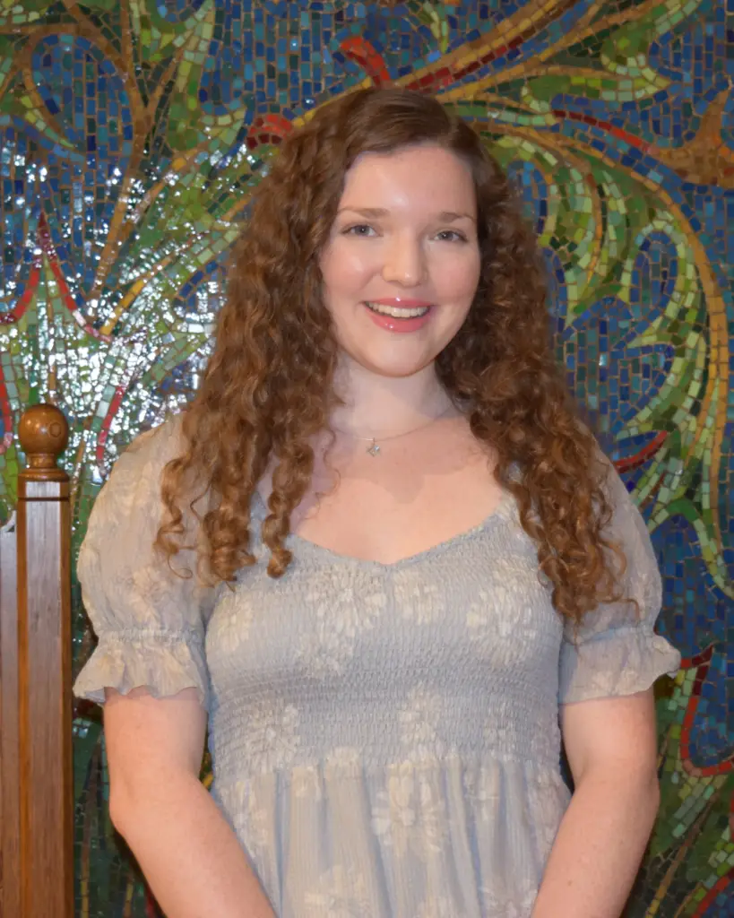 A young woman with curly hair in a light dress smiles while standing before a vibrant mosaic wall at Temple Emanu-El.