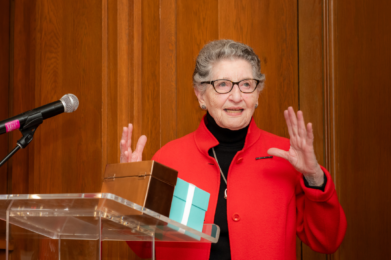 A woman in a red jacket speaks at Temple Emanu-El podium with a gift box, smiling and gesturing warmly with her hands.