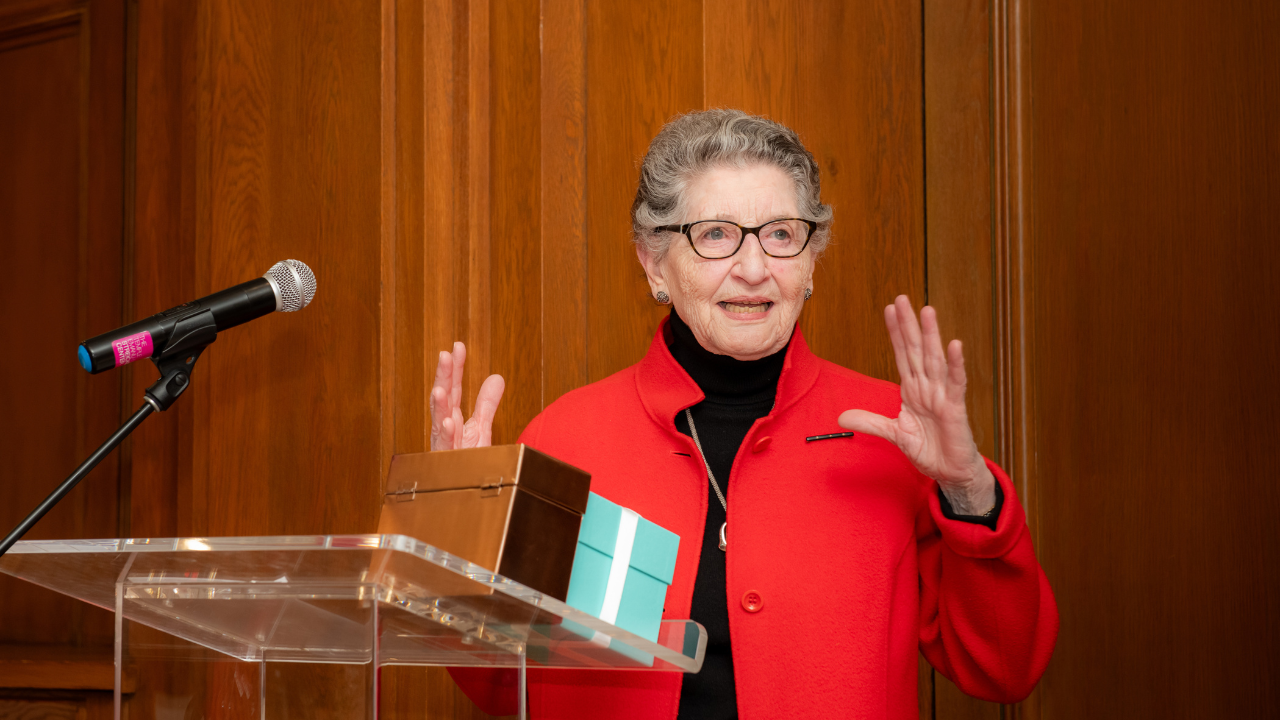 A woman in a red jacket speaks at Temple Emanu-El podium with a gift box, smiling and gesturing warmly with her hands.