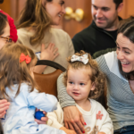 Adults and young children gather with smiles and conversation in a welcoming space at Temple Emanu-El.